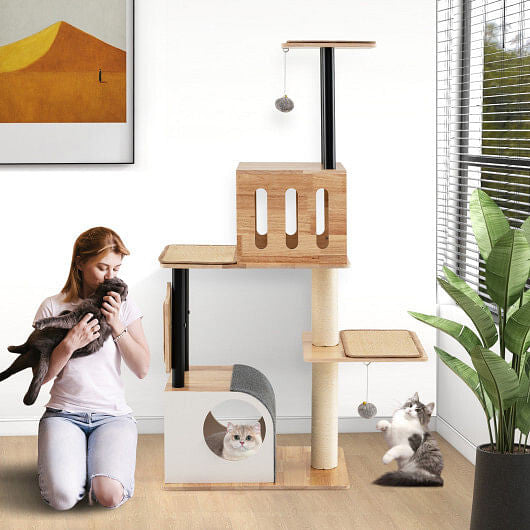 Woman playing with a cat next to a wooden cat tree in a room with a plant and window.